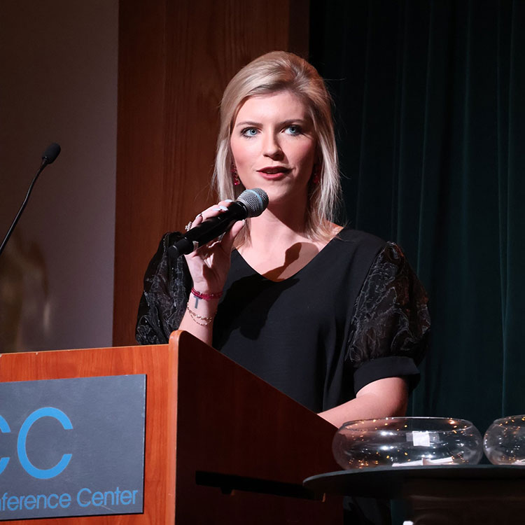 A woman with blonde hair holding a microphone and standing behind a podium talking, wearing a black shirt.